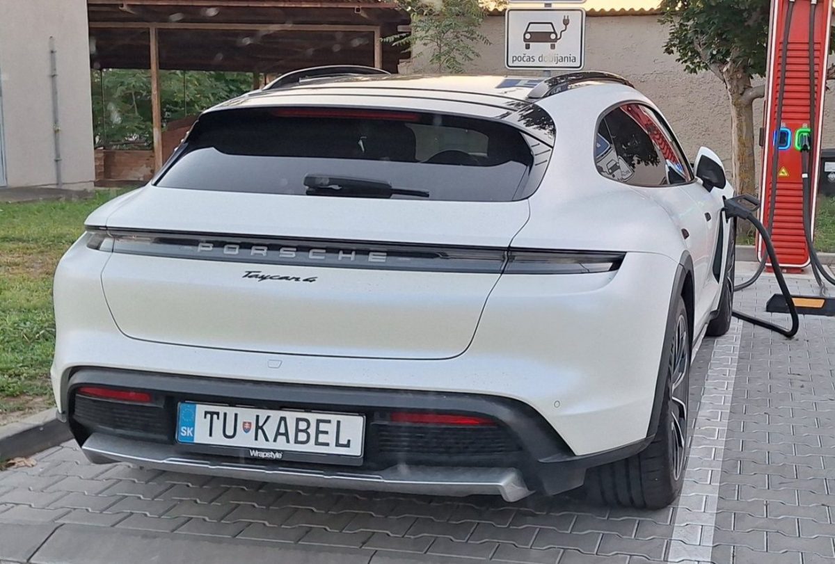 Biele Porsche Taycan 4 nabíjajúce sa na nabíjacej stanici s jedinečnou poznávacou značkou.