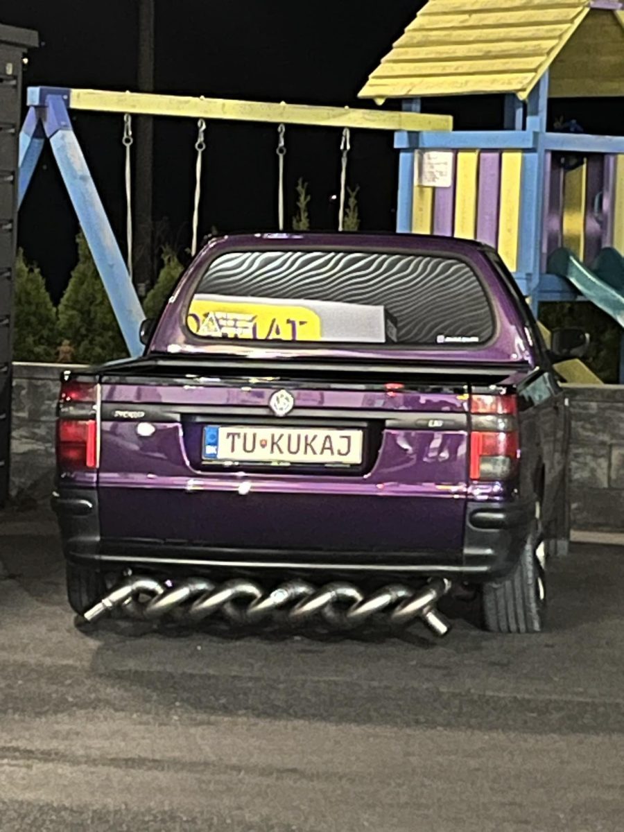 Fialové auto s originálnym výfukom a značkou TU KUKAJ zaparkované vedľa detského ihriska v noci.