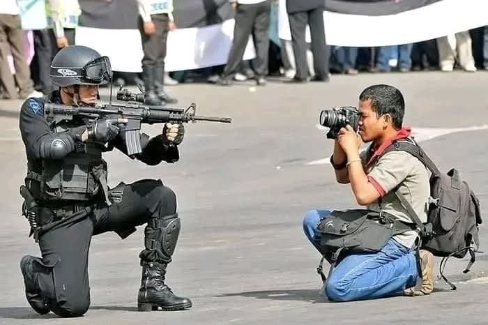 Fotograf kľačí a fotí policajta s puškou počas udalosti na ulici.