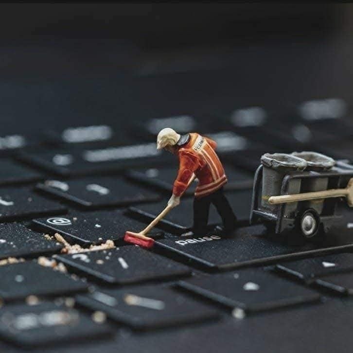 Miniatúrny pracovník čistí klávesnicu počítača s metlou a vozíkom na odpad. Kreatívna scéna z každodenného života.