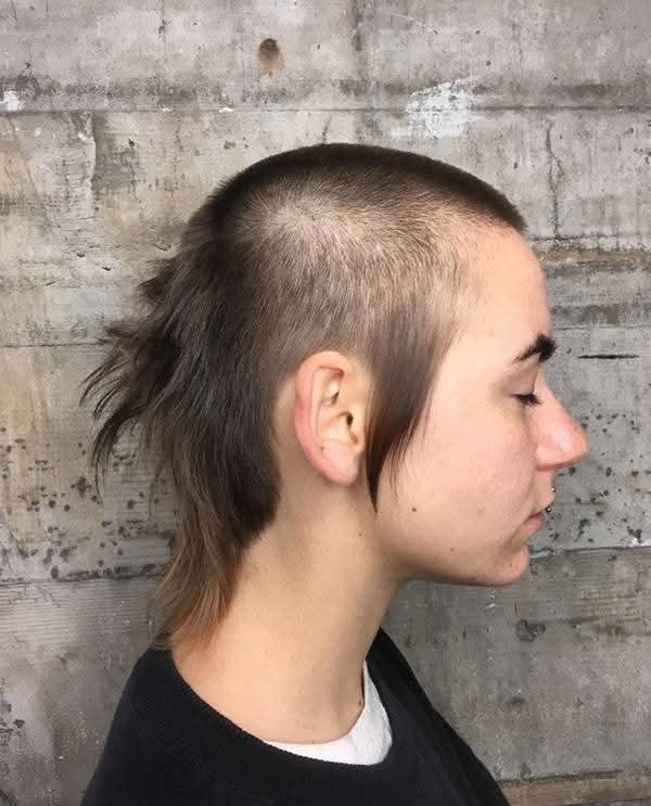 Moderný účes s vyholenými stranami a dlhšími vlasmi vzadu na šedom pozadí.