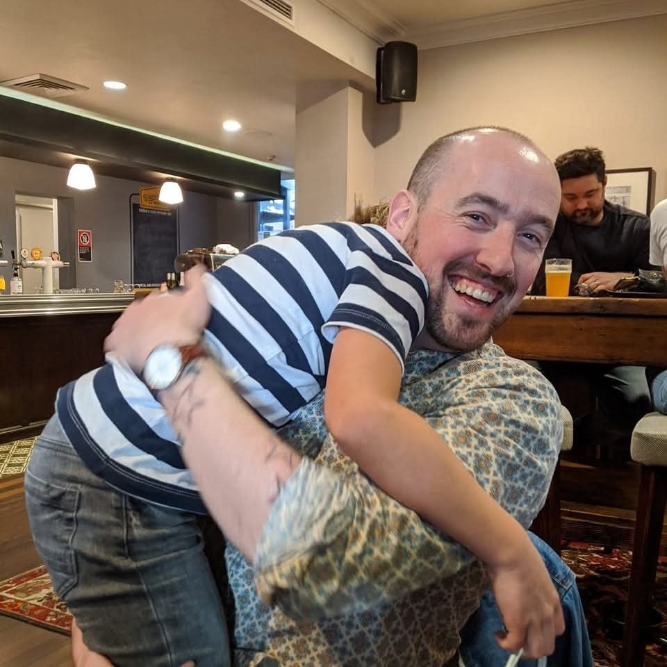 Usmievajúci sa muž objíma ďalšieho človeka v interiéri.