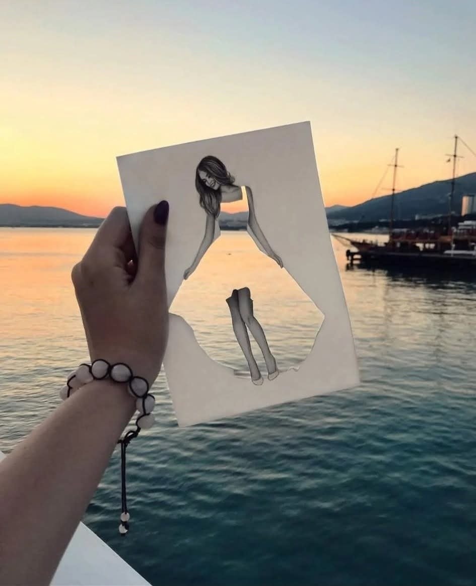 Ruka drží papier s vystrihnutou siluetou pred západom slnka nad vodou.