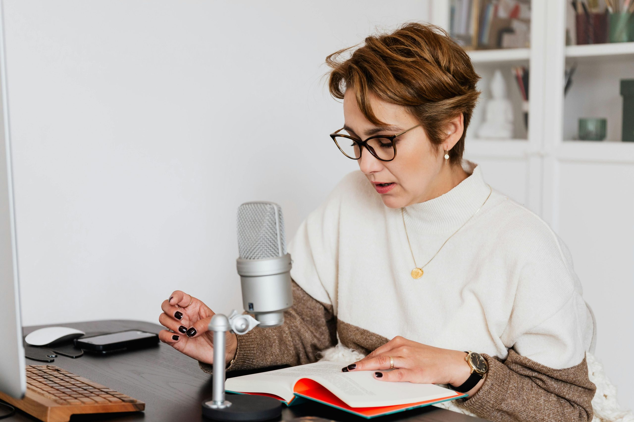Žena s okuliarmi a mikrofónom nahráva podcast pri čítaní knihy v kancelárii.