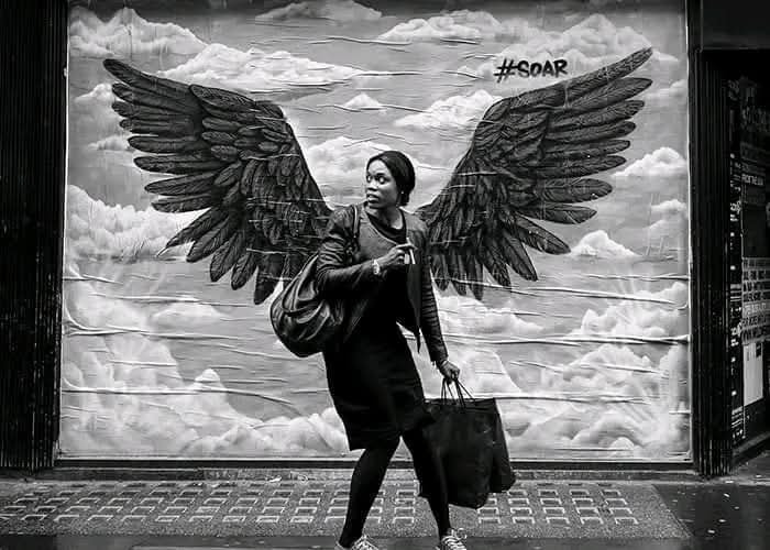 Žena kráča pred nástennou maľbou anjelských krídel, hashtag #SOAR. Čiernobiela fotografia s dynamickou kompozíciou.