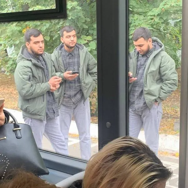 Trojica mužov v rovnakom oblečení čakajúci na autobus pri ceste, držiac telefóny, s odrazmi v okne.