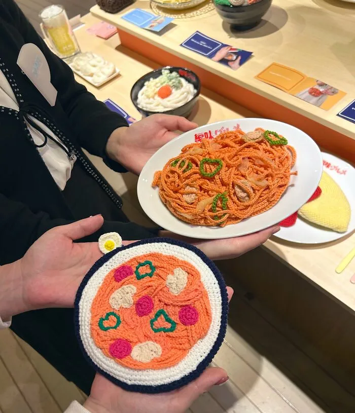 Dve ruky držia háčkované jedlá, napodobňujúce špagety a pizzu, vystavené na pultíku v reštaurácii.