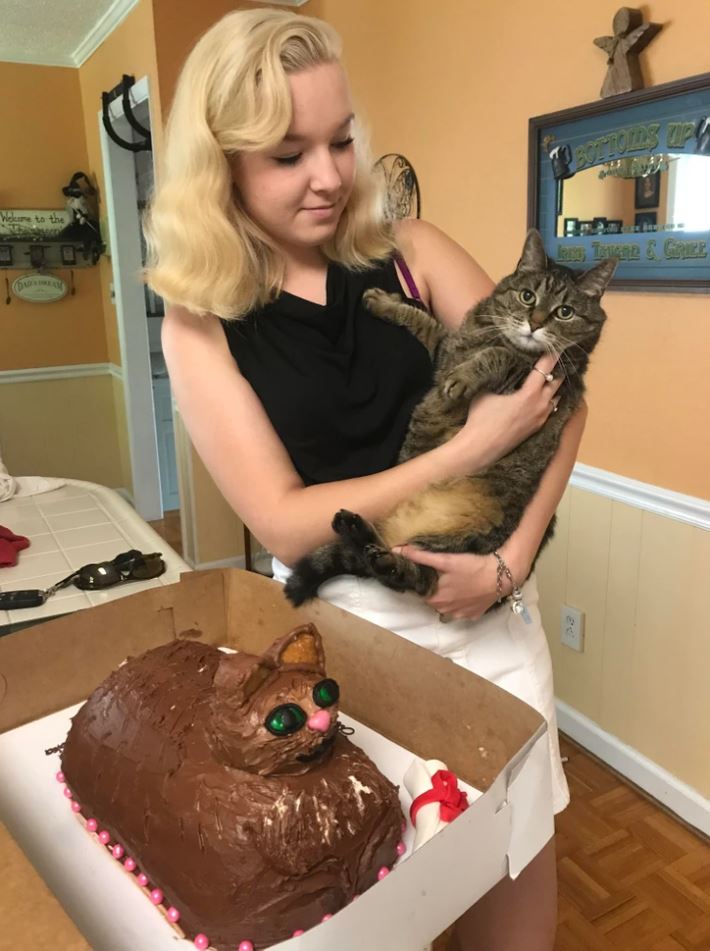 Žena drží mačku pri torte v tvare mačky, ozdobenej čokoládovou polevou a farebnými detailmi.