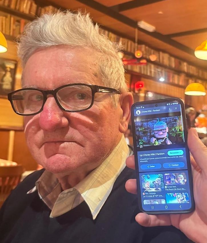 Muž v kaviarni, porovnávanie s postavou na obrázku v mobile, plná knižnica v pozadí.