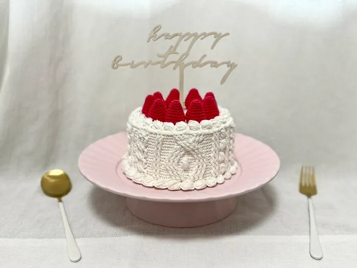 Háčkovaná torta s nápisom happy birthday a dekoráciou z červených háčkovaných bobúľ na ružovom tanieri.