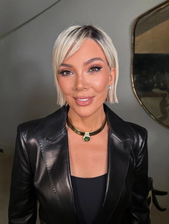 Žena s krátkymi blond vlasmi v čiernej koženej bunde a náhrdelníku so zeleným kameňom, usmievajúca sa do kamery.