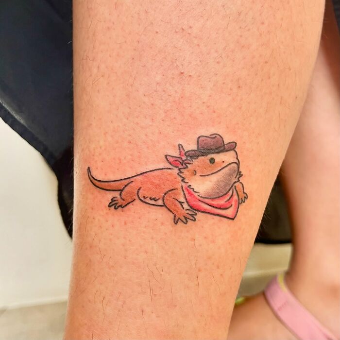 Tetovanie leguána s klobúkom a šatkou na nohe, karikatúrny štýl, farebné.