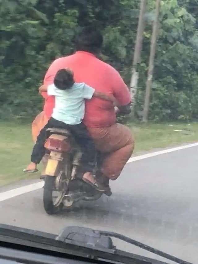 Otec a dieťa na motorke, dieťa objíma otca, jazda po vidieckej ceste s bujnou vegetáciou na pozadí.
