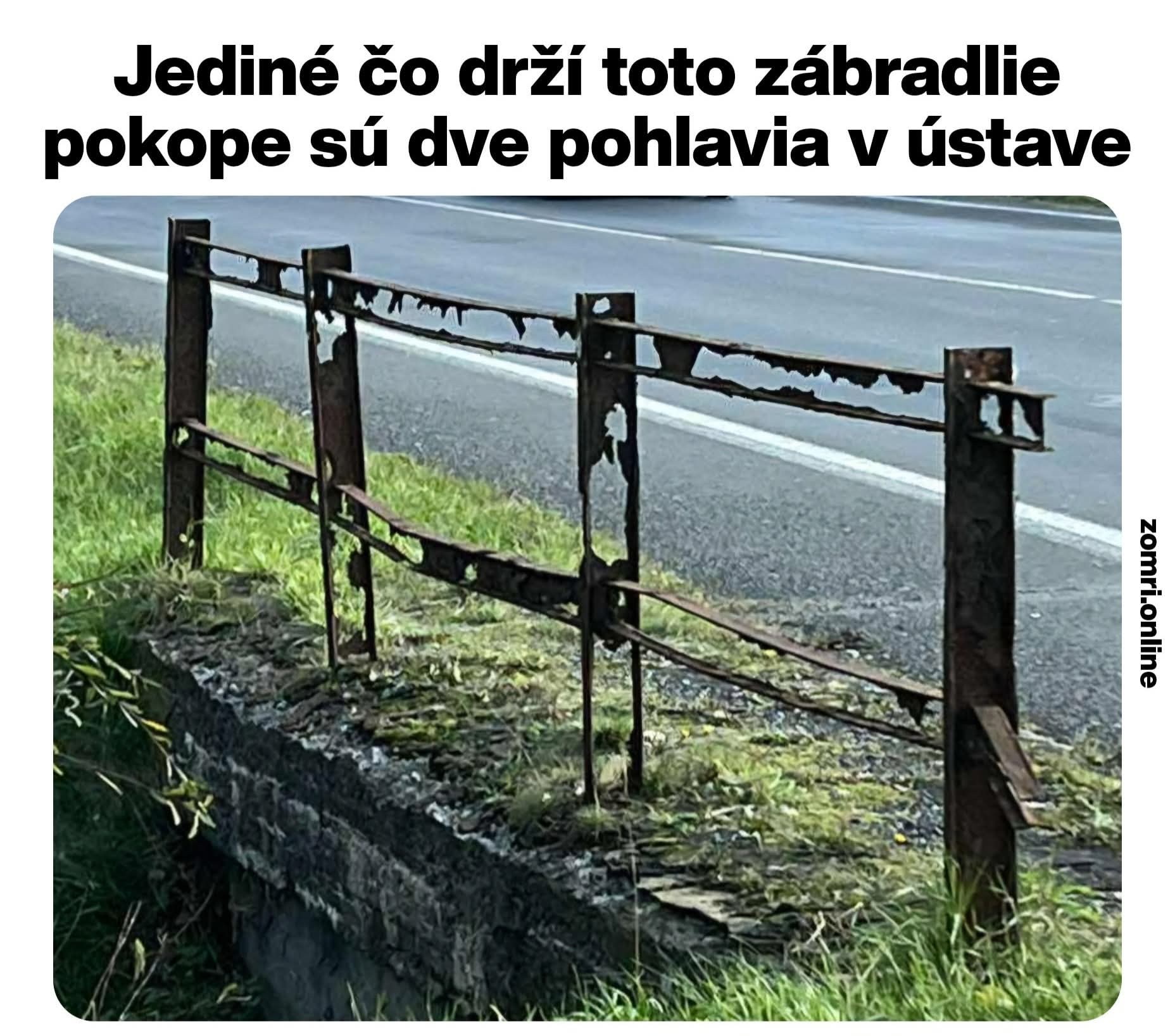 Hrdzavé zábradlie vedľa cesty s poškodenou konštrukciou a trávou na okraji.