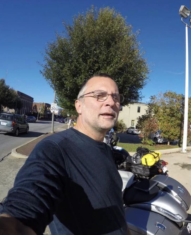 Osoba na ulici vedľa motorky, jasný slnečný deň, strom a budovy v pozadí.