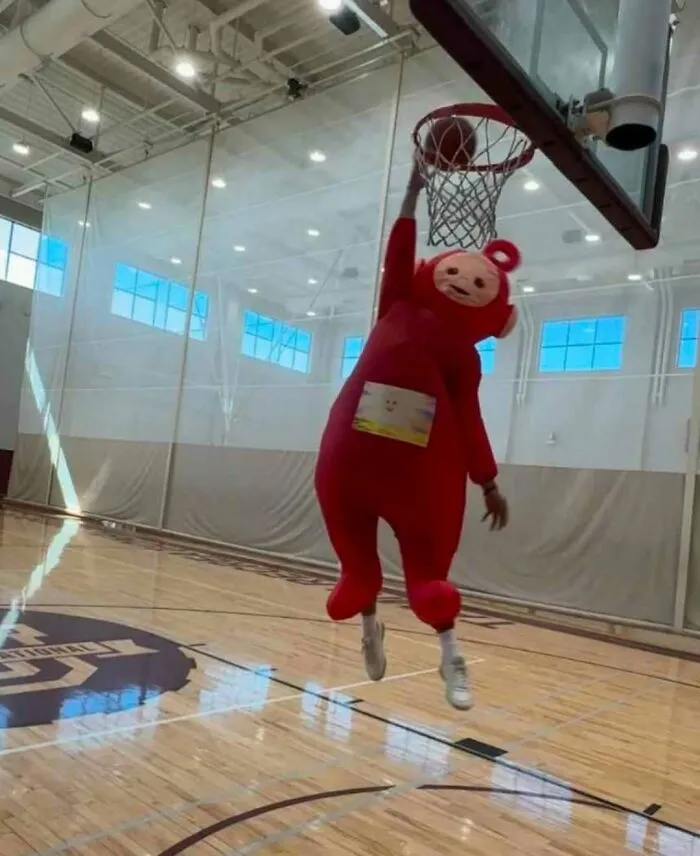 Osoba v červenom kostýme skáče na basketbalovom ihrisku s rukou na koši.