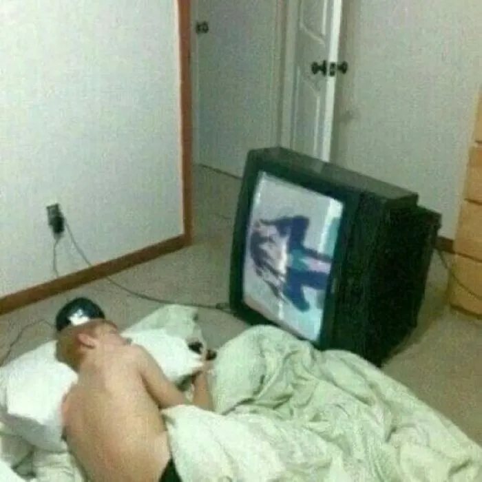 Muž spí na podlahe vedľa televízora, ktorý leží na boku, v neúhľadnej miestnosti.