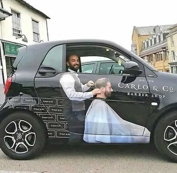 Auto so vtipnou reklamou na holičstvo, kde vodič vyzerá ako holič strihajúci zákazníka na polepe.
