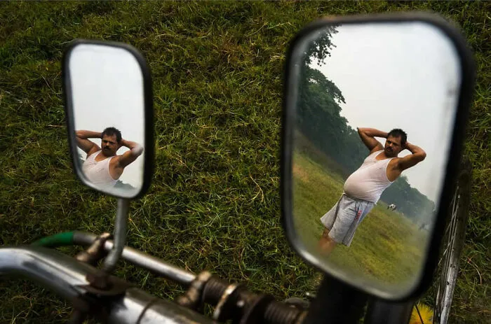 Muž v bielom tričku sa pozerá do spätných zrkadiel na bicykli, stojí na zelenej lúke s rukami za hlavou.
