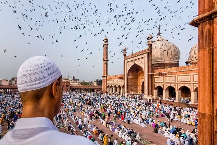 Muslimovia sa modlia pri veľkej mešite, obloha plná vtákov, architektonická krása v pozadí.