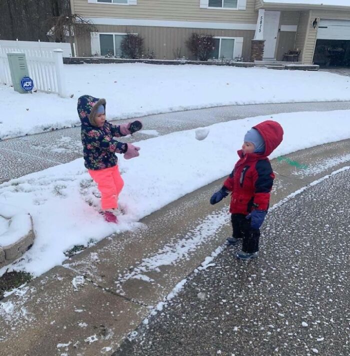 Dve deti sa hrajú so snehom na zasneženej ulici pred domom, jedno z nich hádže snehovú guľu.