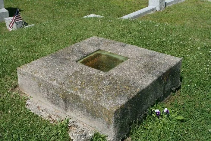 Kamenný pamätník na cintoríne s americkou vlajkou, skleneným štvorcom a fialovým kvetom na tráve.