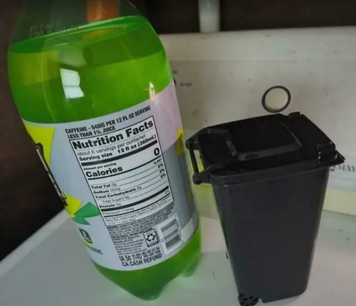 Plastová fľaša limonády vedľa malého odpadkového koša na bielej podlahe.