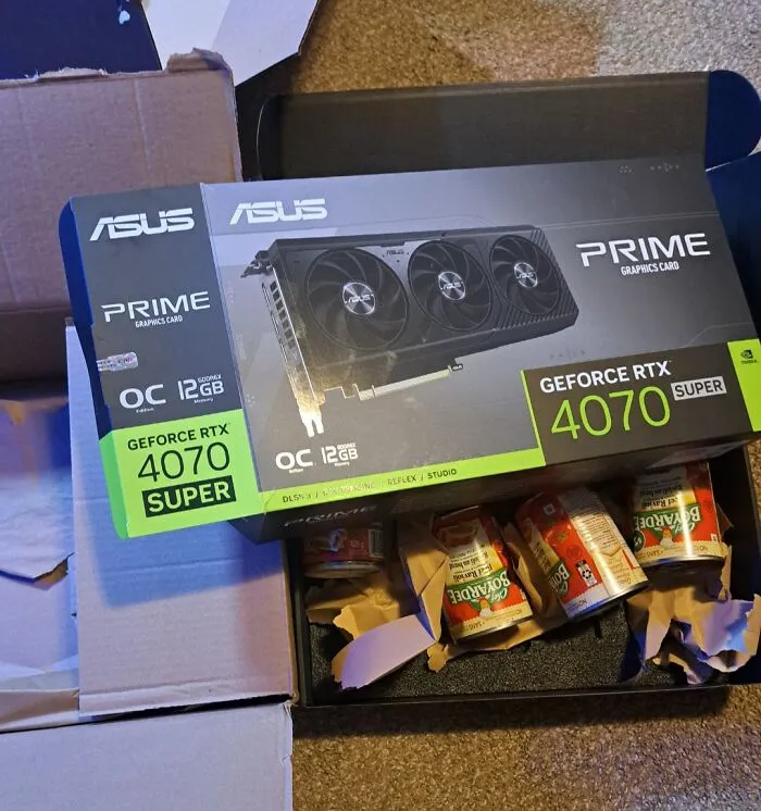 Krabica s grafickou kartou ASUS GeForce RTX 4070 vedľa plechoviek fazule na podlahe.
