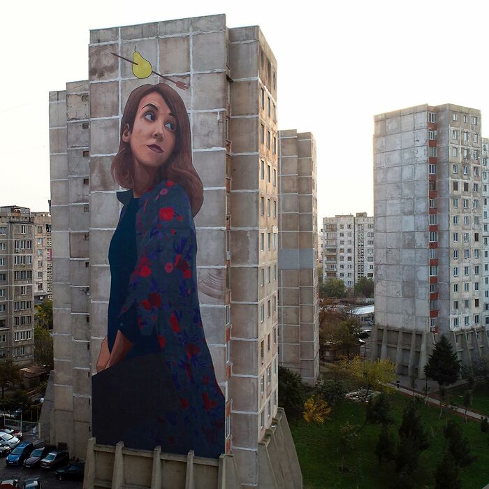 Mural na paneláku zobrazuje ženu s hruškou na streche, obklopenú mestskými domami.