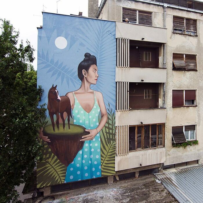 Mural s dievčaťom držiacim koňa na ostrove, maľovaný na stene budovy, s modrou oblohou a rastlinami v pozadí.