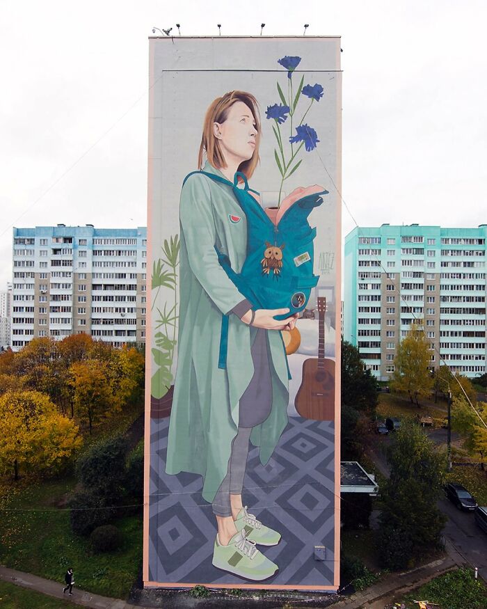 Mural ženy s rastlinami držiace modrú tašku na vysokej budove, obklopený mestskou scenériou.