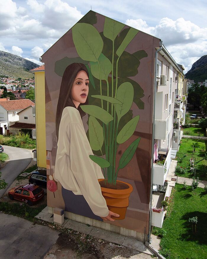 Nástenná maľba ženy s rastlinou na boku budovy, modrá obloha a okolitá krajina.