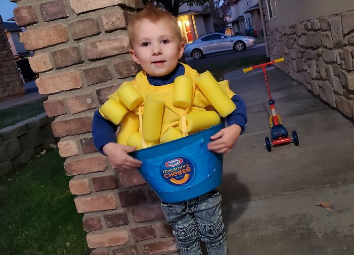 Dieťa v kostýme makarónov a syra s vedrom na ulici vedľa kolobežky.