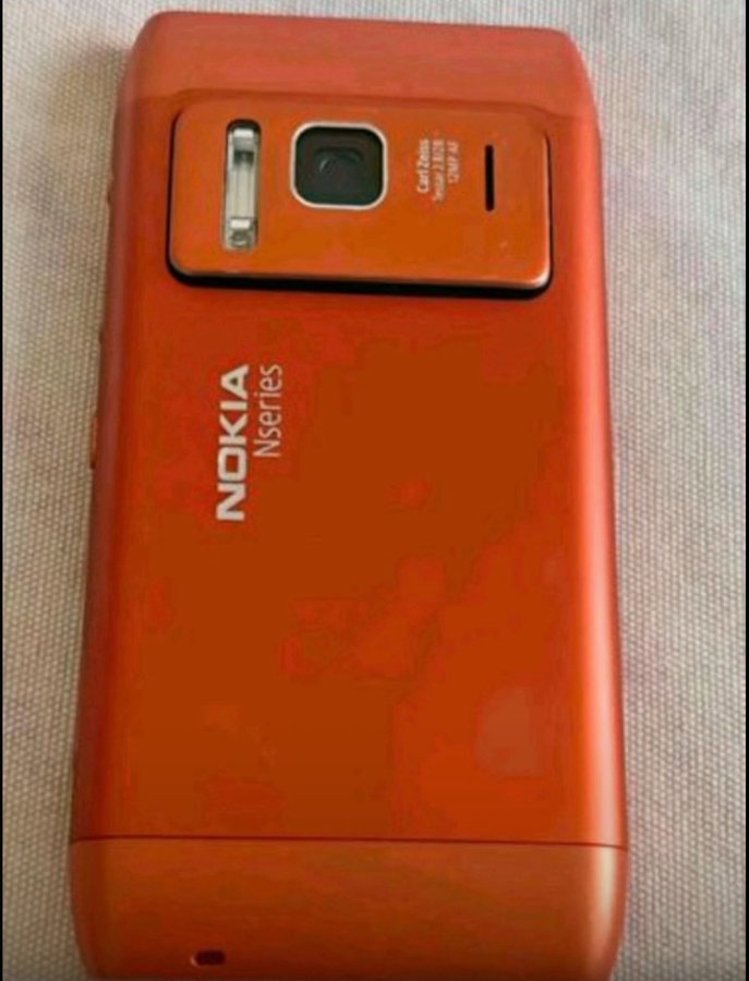 Starý oranžový mobilný telefón Nokia Nseries so zadnou kamerou, položený na bielej ploche.