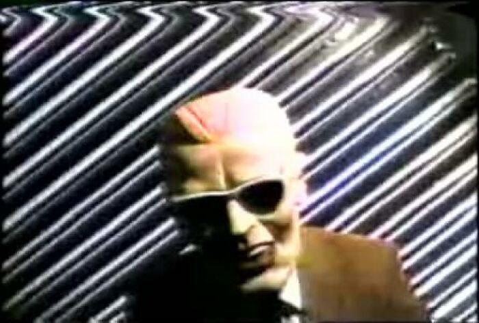 Osoba v maske s okuliarmi pred pruhovaným pozadím v retro štýle.