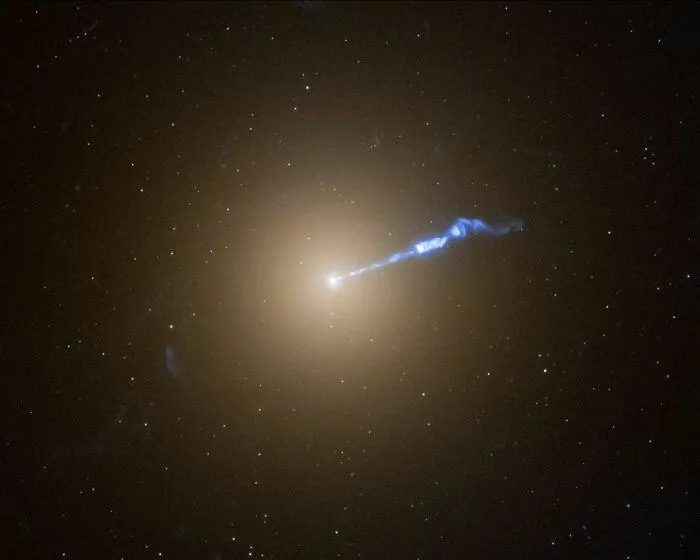 Jasná oranžová galaxia so silným modrým výtryskom svetla na tmavom vesmírnom pozadí plnom hviezd.