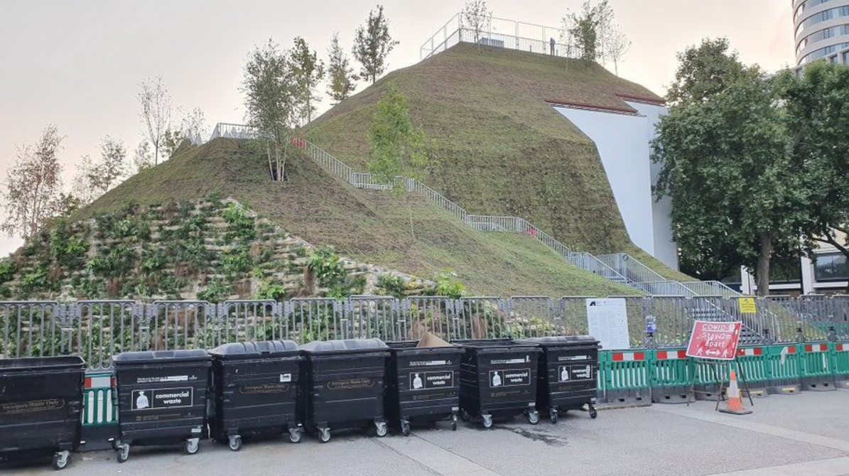 Umelý kopec v mestskom prostredí s kontajnermi na odpad v popredí, zobrazuje zeleň a architektúru.