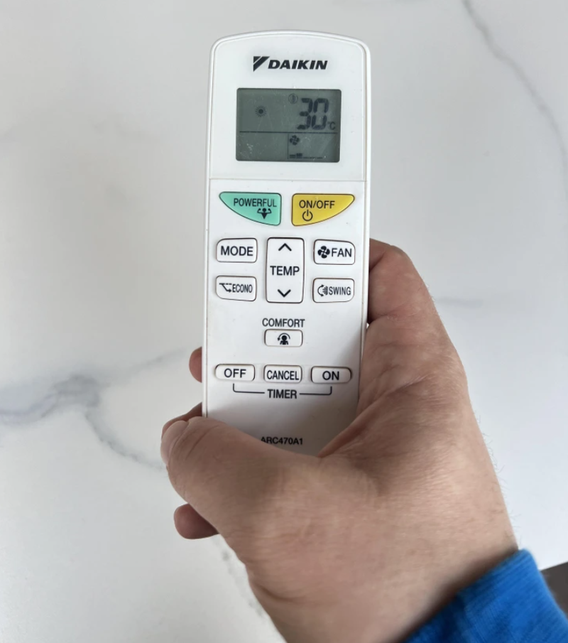 Diaľkový ovládač klimatizácie Daikin s nastavenou teplotou 30°C, držený v ruke na bielom pozadí.