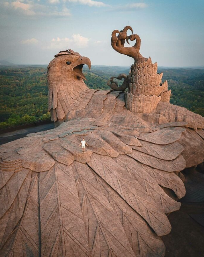 Obrovská kamenná socha vtáka v prírode, s osobou stojacou na jeho rozsiahlom tele.