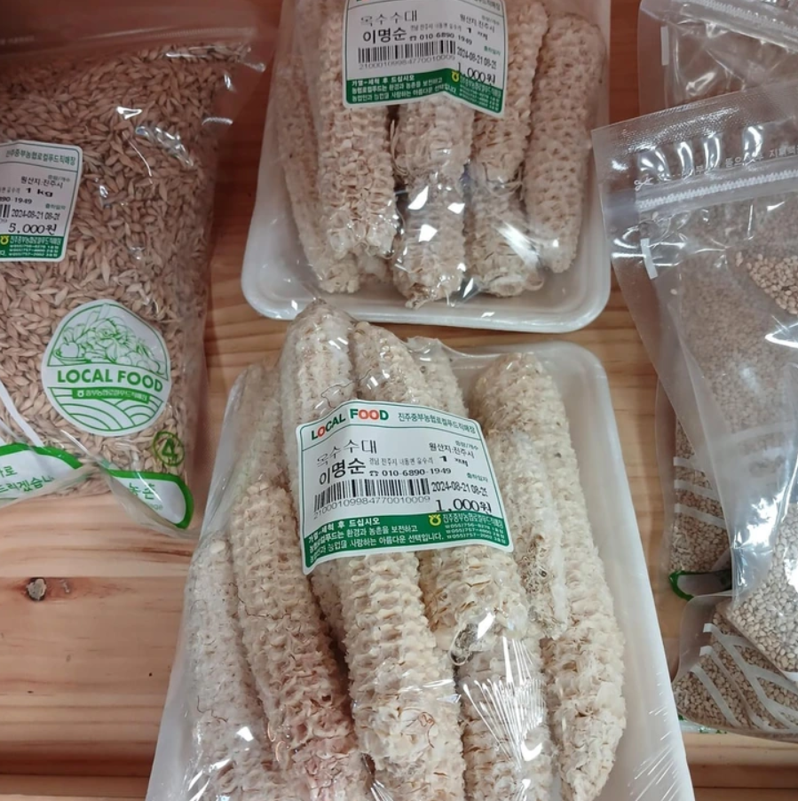 Balíky obilnín a sušených kukuričných šúľkov na drevenom stole, označené ako miestne potraviny.