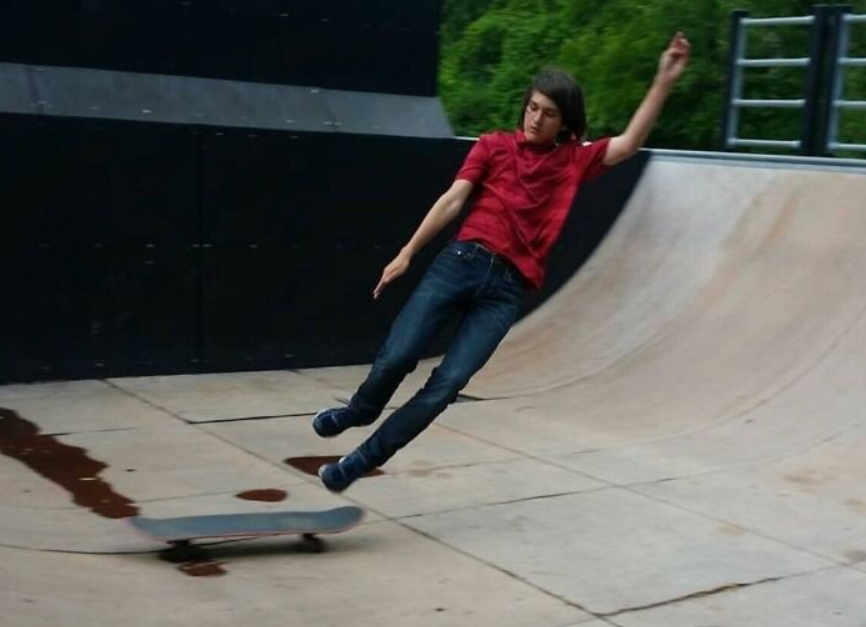 Chlapec padá zo skateboardu v skateparku, oblečený v červenej košeli a džínsoch, so zeleným pozadím.