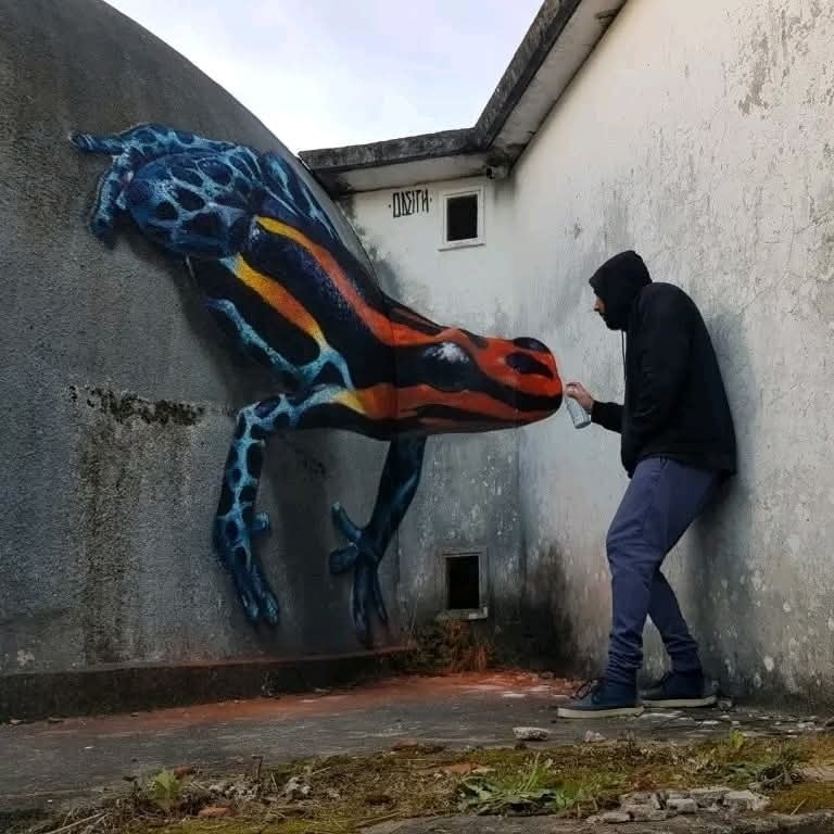 Umelec strieka dúhovú jaštericu na stenu, vytvára realistický 3D street art v betónovej uličke.