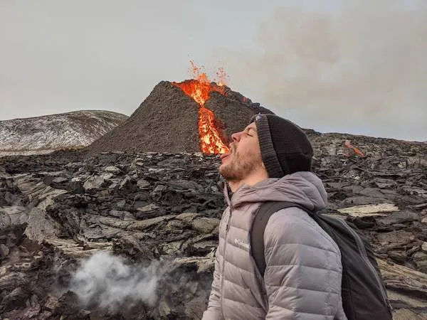 Turista žartovne „požiera“ lávu z erupcie sopky v pozadí, oblečený v zime na kamenistej kraji.