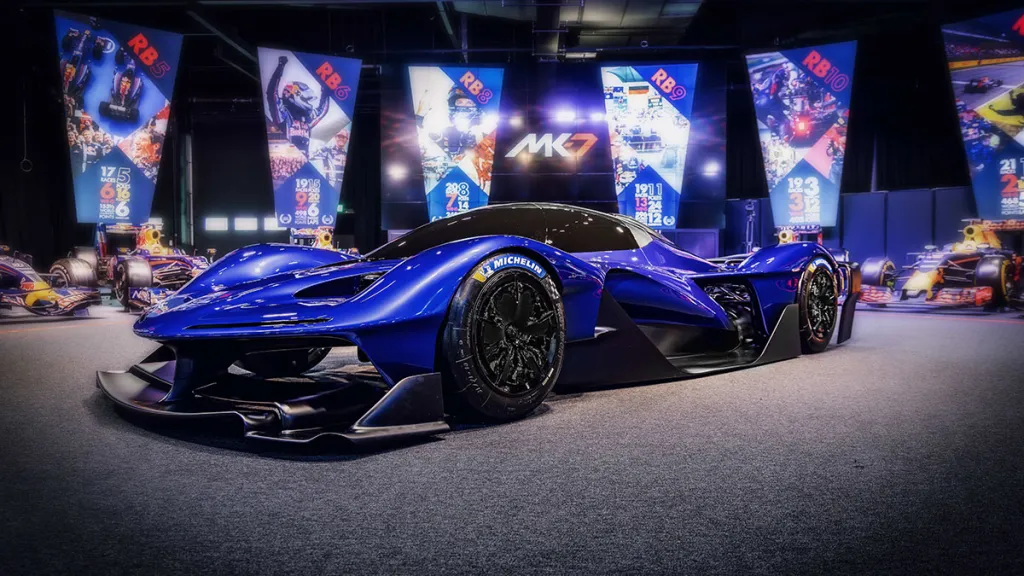 Modrý superšportiak v štýle futuristického pretekárskeho auta vystavený na autosalóne, obklopený farebnými bannermi.