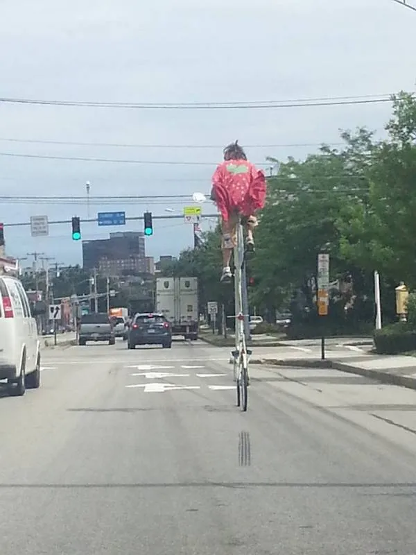 Osoba v červenej bunde na vysokom bicykli jazdí po mestskom bulvári plnom áut.