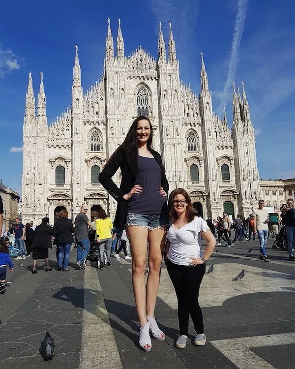 Dve ženy pred milánskou katedrálou, výrazný kontrast výšky, modrá obloha, turistická atrakcia.