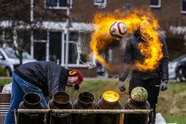 Muž odpaluje horiacu futbalovú loptu z kovového kanistru v exteriéri, vytvárajúci dramatický efekt ohňa.