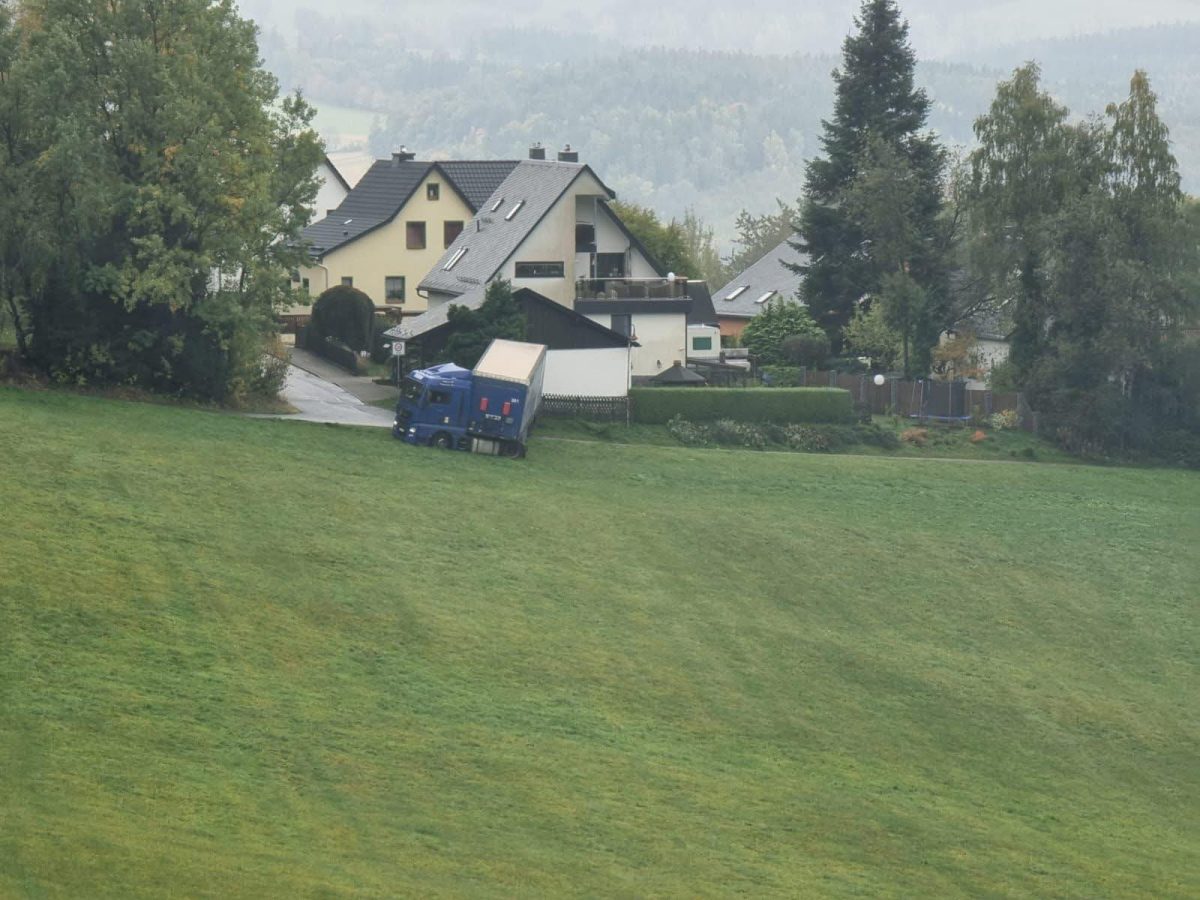 Kamión stojaci na svahu pred domami v pokojnej vidieckej oblasti, obklopený zeleňou a stromami.