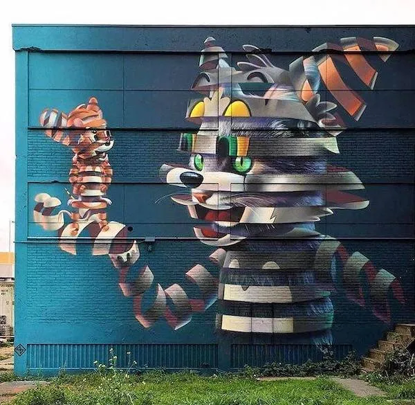 Street art mural s farebným 3D efektom predstavujúci pruhovanú mačku s mláďaťom na modrom pozadí budovy.