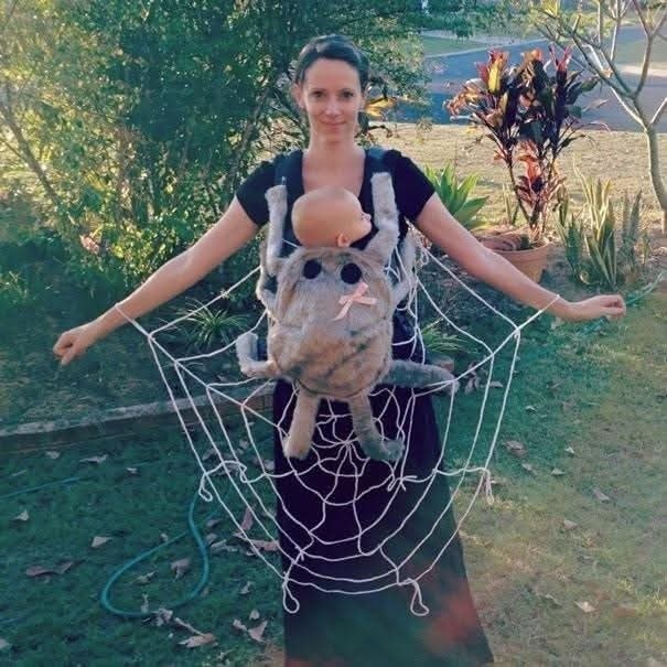 Žena v kostýme pavúka s bábätkom v nosiči, obaja oblečení do pavučiny na záhrade.